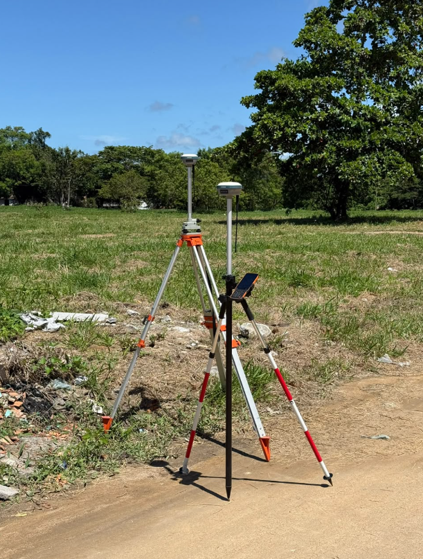 Profissional em campo com receptor GNSS e bastão em estrada de acesso.